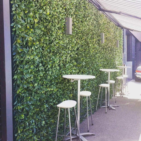 An outdoor patio featuring Artificial Laurel Hedge Plant Wall Screening Panels, gray wall sconces, white-topped bar tables with stools, and a partial awning in a seating area