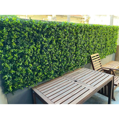 An outdoor patio with Artificial Spring Sensation Hedge Wall Panels installed as the backdrop to a wooden table and chair in the seating area